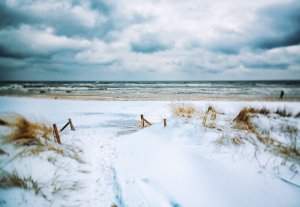 A snowy beach scene with rolling sand dunes covered in snow leading to a gray, wavy ocean. The sky is overcast with thick clouds, adding to the chilly atmosphere. Sparse dry grass pokes through the snow near the wooden path.