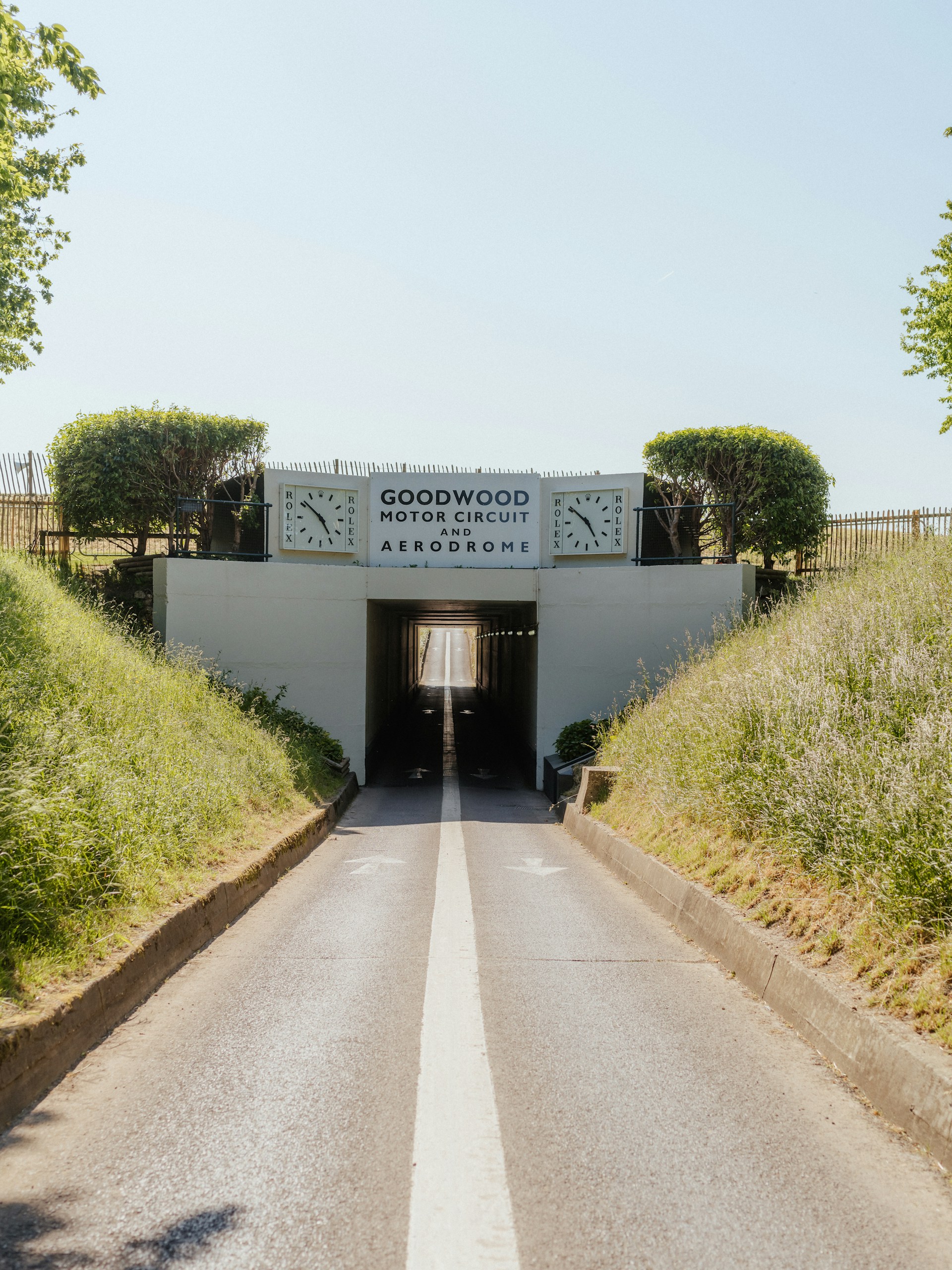 A road winds through a grassy embankment, passing under a tunnel marked Goodwood Motor Circuit Aerodrome, with two clocks on the sign and trees above—one of the Top Things to Do in West Sussex When the Sun is Shining.